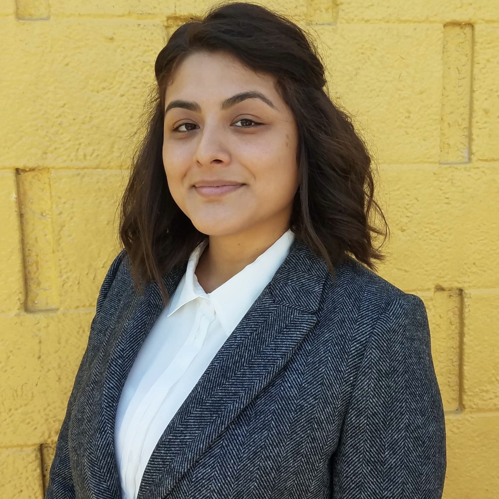 Woman with dark hair wearing jacket and white shirt in front of a yellow wall