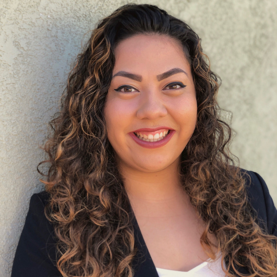 Woman with brown curly hair wearing a white shirt and black suit jacket smiling for a photo