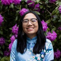 woman in blue floral shirt and glasses smiles brightly in front of plant with purple flowers