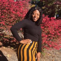 woman poses outside in front of red flowers. She is wearing striped pants and a black shirt