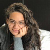 woman with glasses rests her head on her hand, looking into the camera conversationally